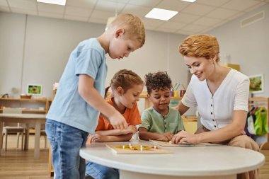 Gülümseyen öğretmen ve ırklar arası çocuklar Montessori okulunda sınıfta didactic materyallerle oynuyorlar.