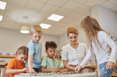 Montessori okulunda oynayan çok ırklı çocukların yanında oturan öğretmen.