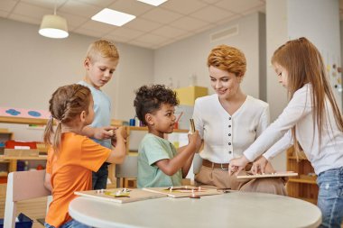 tebessüm eden çoklu etnik çocuklar Montessori okulunda öğretmenin yanında didactic materyaller tutuyorlar.