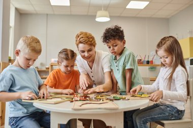 Gülümseyen öğretmen Montessori okulunda oyun masasında çoklu etnik kökenli çocuklarla oynuyor.