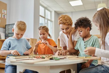 Montessori okulunda sınıf öğretmeninin yanında oyun oynayan ırklar arası çocuklar.