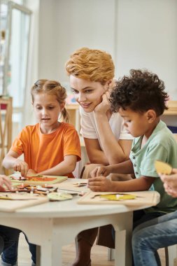 Gülümseyen öğretmen Montessori okulunda çok ırklı çocukların yanında oturmuş didactic materyallerle oynuyor.