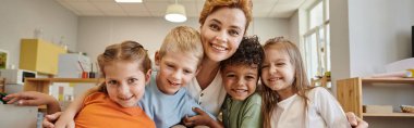 Neşeli öğretmen ve ırklar arası çocuklar Montessori dersi sırasında kameraya bakıyor.