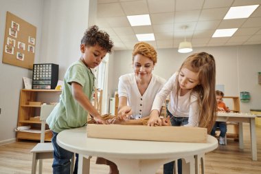 Gülümseyen öğretmen ve çoklu etnik çocuklar Montessori okulunda ders sırasında tahta çubuklar alıyorlar.