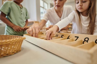 Montessori sınıfındaki çoklu etnik çocukların yanında tahta sopa yiyen öğretmen manzarası.