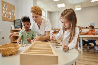Öğretmen Montessori okulunda tahta çubuklarla oynarken çok ırklı çocuklarla konuşuyor.