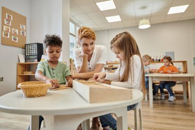 Öğretmen Montessori okulunda ders sırasında tahta sopalarla oynayan çoklu etnik kökenli çocukların yanında oturuyor.