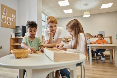 teacher sitting near multiethnic kids playing with wooden sticks during lesson in montessori school clipart