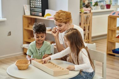 Öğretmen Montessori okulunda konuşurken gülen çoklu etnik çocuklar tahta sopalarla oynuyorlar.