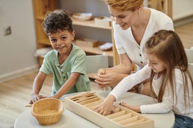 Mutlu Afro-Amerikalı çocuk Montessori sınıfında öğretmen ve arkadaşının yanında oynuyor ve kameraya bakıyor.