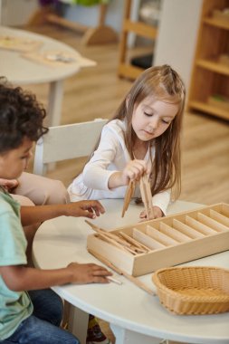 Montessori okulunda ders sırasında masada tahta sopalarla oynayan ırklar arası çocuklar.