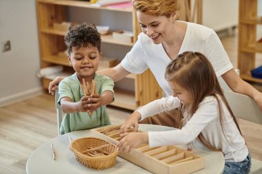 Montessori okulunda öğretmenlerin yanında ahşap didactic malzemelerle oynayan pozitif çoklu etnik çocuklar