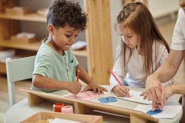 Montessori okulunda öğretmenlerin yanında kalemle çizim yapan çok ırklı çocuklar.