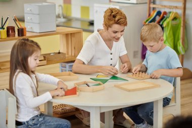Gülümseyen öğretmen çocuklarla oynuyor ve Montessori okulunda masada didactic malzemeler var.