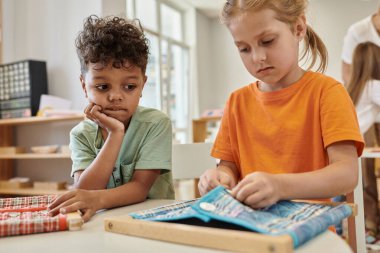 Afrikalı Amerikalı çocuk Montessori okulunda kumaş ve düğmelerle oynayan arkadaşına bakıyor.