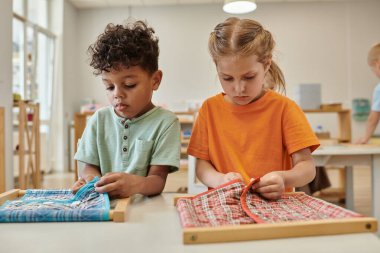 Montessori okulunda sınıftaki çok ırklı çocuklar kumaşları çerçevelere iliştiriyor.