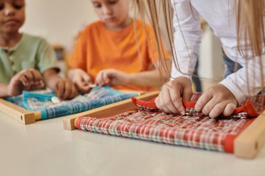 Montessori okulunda çoklu etnik arkadaşların yanında kumaş ve düğmelerle oynayan çocuk manzarası