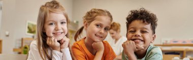 smiling and interracial kids looking at camera in blurred class in montessori school, banner clipart