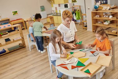 Gülümseyen öğretmen Montessori okulunda çoklu etnik çocuklarla üçgenlerle oynuyor.