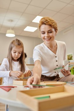 cheerful teacher holding triangles near pupil in classroom in montessori school clipart