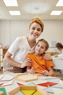 Pozitif öğretmen kucaklayan kız ve Montessori okulunda sınıfta kameraya bakmak.