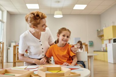 Gülümseyen öğretmen Montessori okulunda didactic materyallerin yanında kızı kucaklıyor.