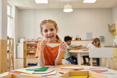 Pozitif çocuk tahta üçgen tutuyor ve Montessori okulunda kameraya bakıyor.