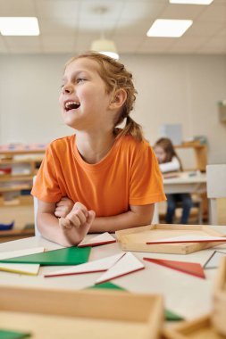 happy child looking away near wooden didactic materials in class of montessori school clipart