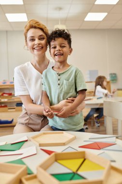 smiling teacher hugging african american kid near didactic materials in montessori school clipart