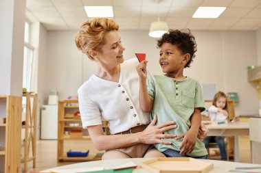 Pozitif Afrikalı Amerikalı çocuk Montessori okulunda ders sırasında öğretmenin yanında üçgen tutuyor.