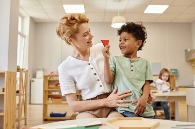positive african american boy holding triangle near teacher during lesson in montessori school clipart