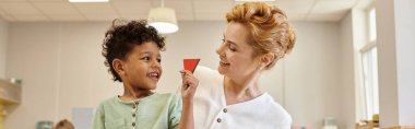 happy african american boy holding triangle near teacher during lesson in montessori school, banner clipart