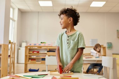 Gülümseyen Afrikalı Amerikalı çocuk bulanık montessori sınıfında dikilirken başka tarafa bakıyor.