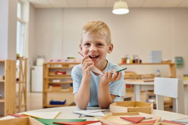 cheerful boy holding triangles during game in classroom in montessori school clipart
