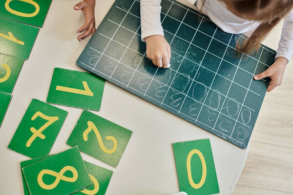 вид сверху девочки, пишущей ноль на доске, которая учится считать в школе Монтессори