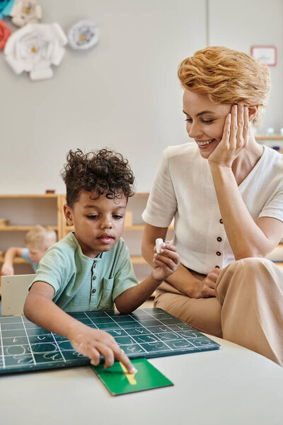 счастливый учитель наблюдает за африканским мальчиком, указывая на число возле доски, учится через игру