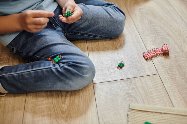 обрезанный вид мальчика, сидящего в джинсах на полу, играя с Монтессори бусы материал, игра