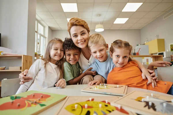 Profesor Sonriente Abrazando Niños Multiétnicos Cerca Materiales Didácticos Mesa Escuela — Foto de Stock