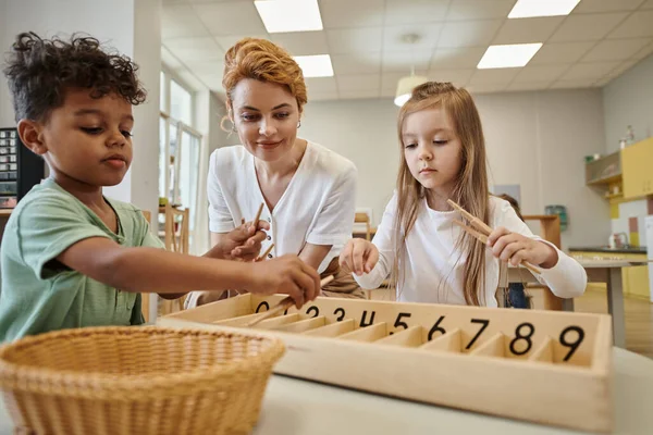 Montessori okulunda gülümseyen öğretmenin yanında tahta sopalarla oynayan çok ırklı çocuklar.