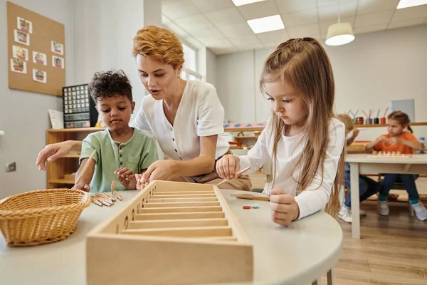 Nauczyciel Rozmawiający Wieloetnicznymi Dziećmi Podczas Zabawy Drewnianymi Kijami Szkole Montessori — Zdjęcie stockowe