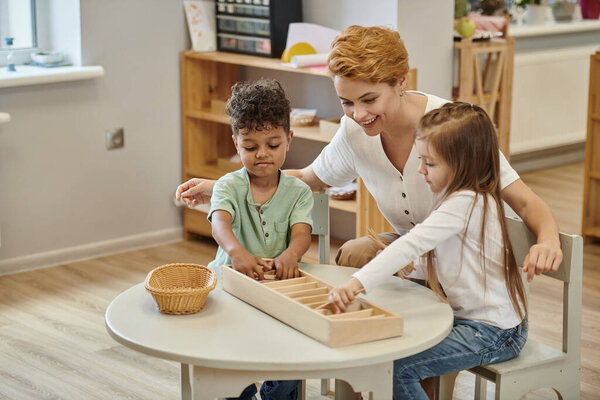 happy teacher talking to interracial kids playing with wooden didactic materials in montessori class
