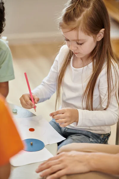 stock image child drawing with pencil near african american friend and teacher in montessori school