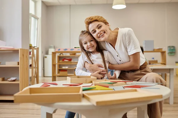 Gülümseyen öğretmen öğrenciye sarılıyor ve Montessori sınıfında kameraya bakıyor.