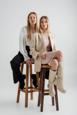 Resmi takım elbiseli iki kız kardeş sandalyede oturup kameraya, moda ve moda kavramına bakıyorlar.
