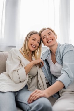 close up two sisters sitting on sofa laughing with closed eyes, hand on knee, family bonding clipart