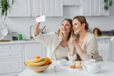 classy sisters in trendy pastel cardigans making selfie at table in kitchen, family bonding clipart