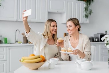 İki modaya uygun kız kardeş selfie çekiyor gülümsüyor ve kek yiyor mutfak zemininde, aile bağı kuruyor.