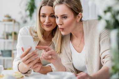 Moda ve şaşırmış kız kardeşler mutfak arka planında cep telefonuna bakıyor, aile bağı kuruyor.
