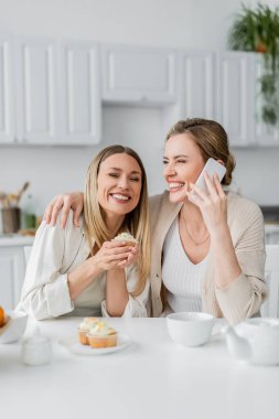 Gülen güzel kız kardeşler kek tadıyorlar ve mutfak arka planında telefon tutuyorlar, aile bağı kuruyorlar.