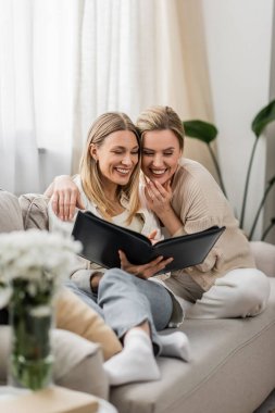 pretty and lovely sisters in trendy attire laughing and looking at photo album, family bonding clipart
